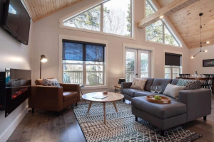 The image shows a cozy, modern living room with a high, vaulted ceiling made of wood paneling. Large triangular windows and glass doors let in natural light and offer a view of trees outside. The space features a gray sectional sofa, a brown leather armchair, and a round coffee table on a patterned rug. A mounted electric fireplace and a wall-mounted TV are visible on the left, while a dining area with a table and chairs is situated in the background. The decor includes neutral tones and minimalist furnishi