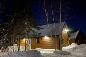 The image shows a cozy chalet at night during winter. The chalet has warm, golden-brown siding, illuminated by exterior lights, creating a welcoming glow. Snow surrounds the building, with tall trees in the foreground and background. The night sky is dark and clear, adding to the serene and peaceful atmosphere.